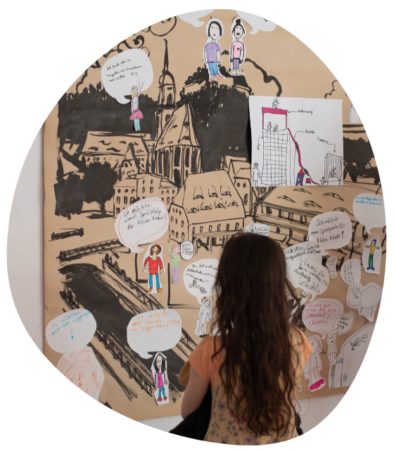 Ein Kind steht vor einer Pinnwand mit einer Zeichnung von Königstein und Sprechblasen mit Wünschen von Kindern für die Stadt.
