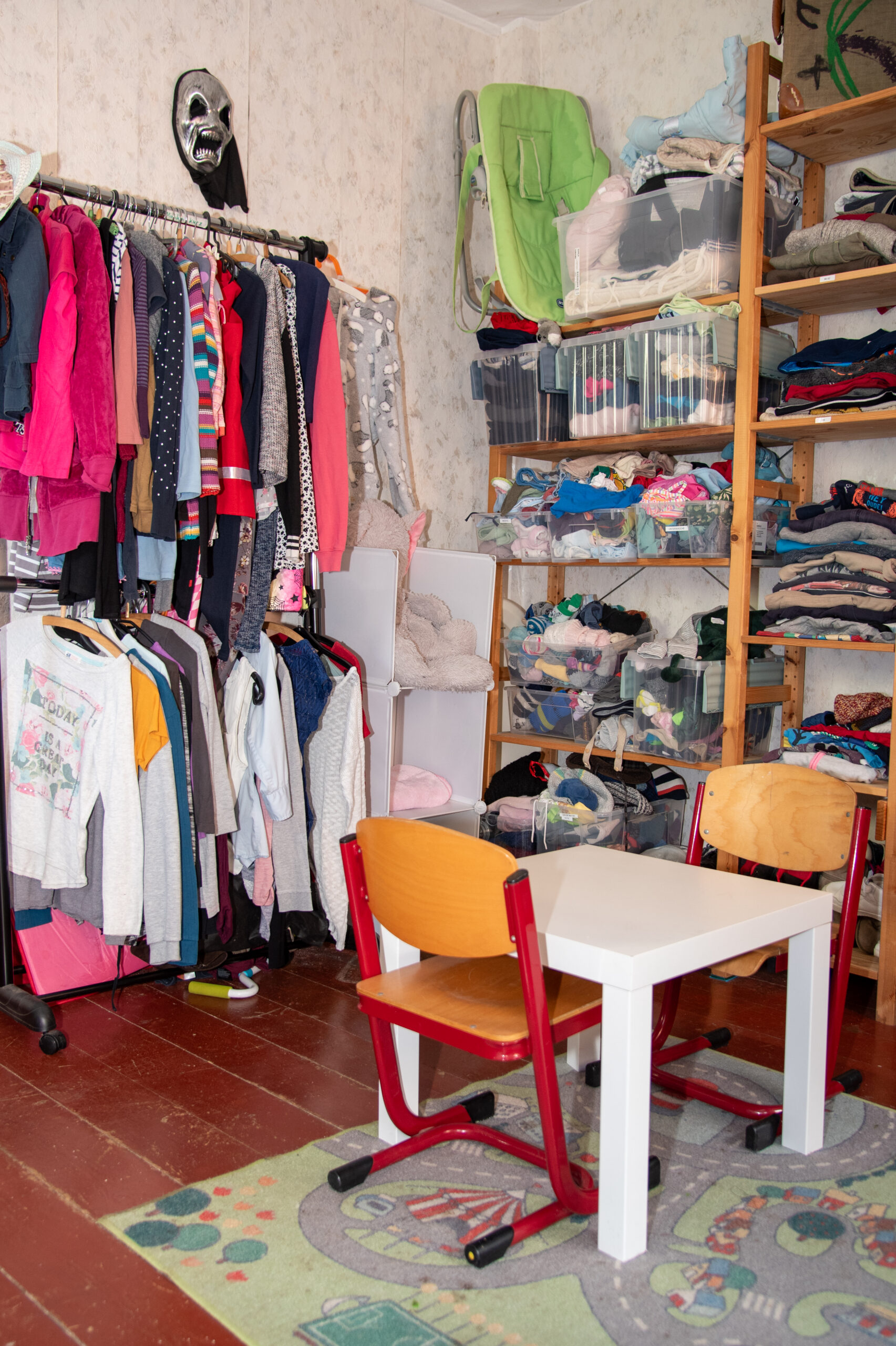 Kinderkleidung. kurzarm und langarm Shirts hängen auf einem Ständer. In einem Holzregal liegen Hosen. im vordergrund steht ein kleiner Tisch mit 2 Stühlen an denen Kinder sitzen und sich beschäftigen können