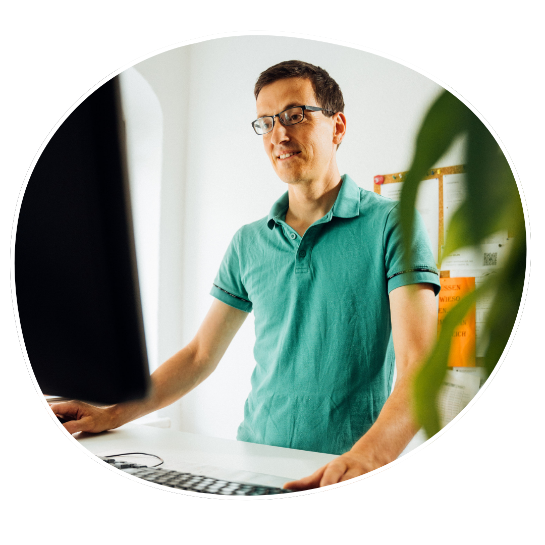 Johannes, ein mann Steht am PC Arbeitsplatz, Trägt ein grünes Shirt. Johannes hat kurzwes dunkles hat und trägt eine Brille.